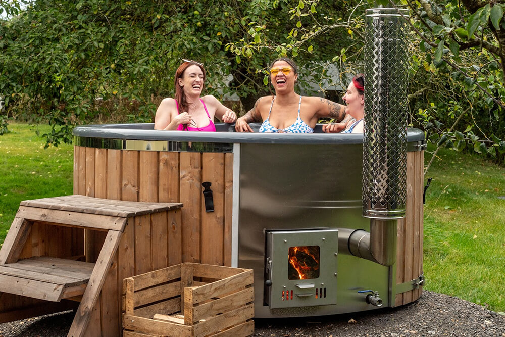 Three girls laughing in a wood fired hot tub surrounded by trees.